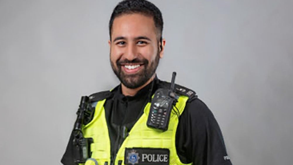 Sergeant Taz Gill in uniform against a white wall, smiling direct to camera. 