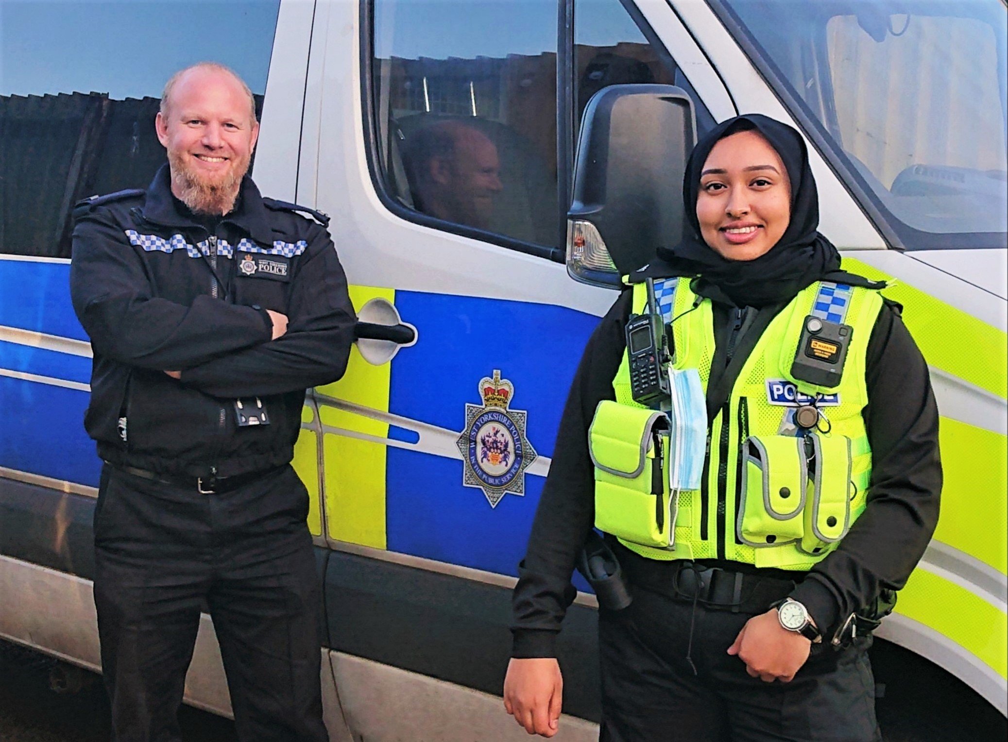 Male and female Muslim officers standing next to a police van and smiling to camera.