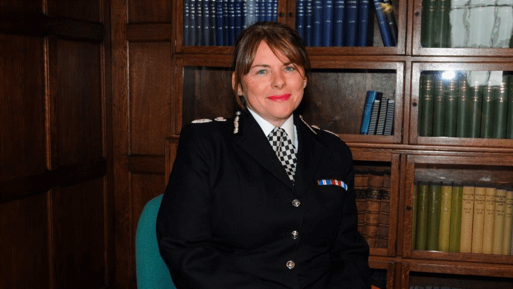 Chief Constable Michelle Skeer in uniform, sitting at a desk, with books on shelves behind her. 