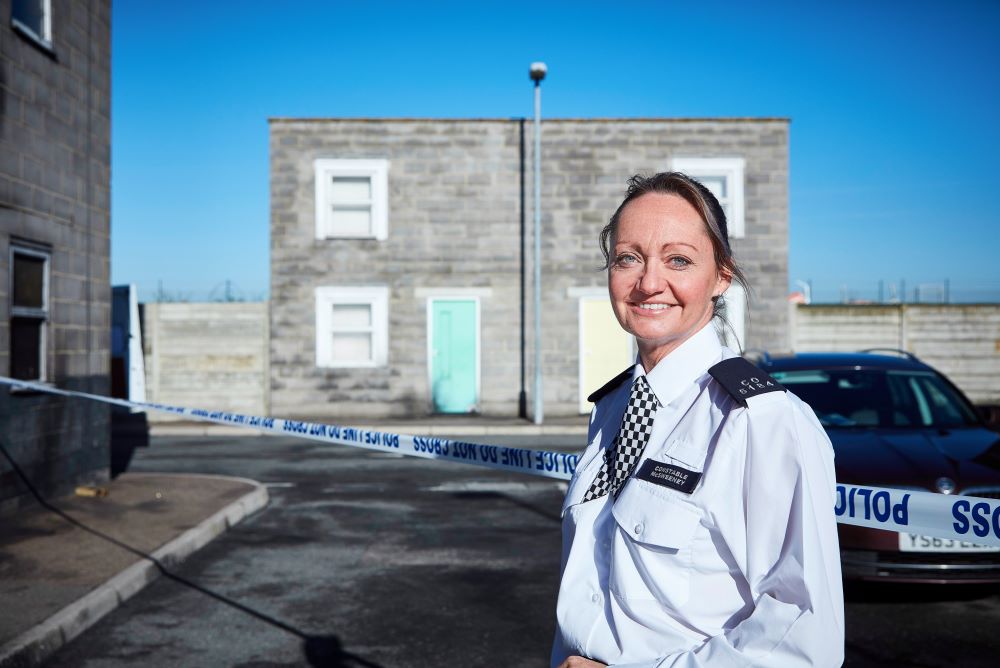 Sergeant Faye McSweeney in uniform, standing outside with police tape, a car and building behind her.