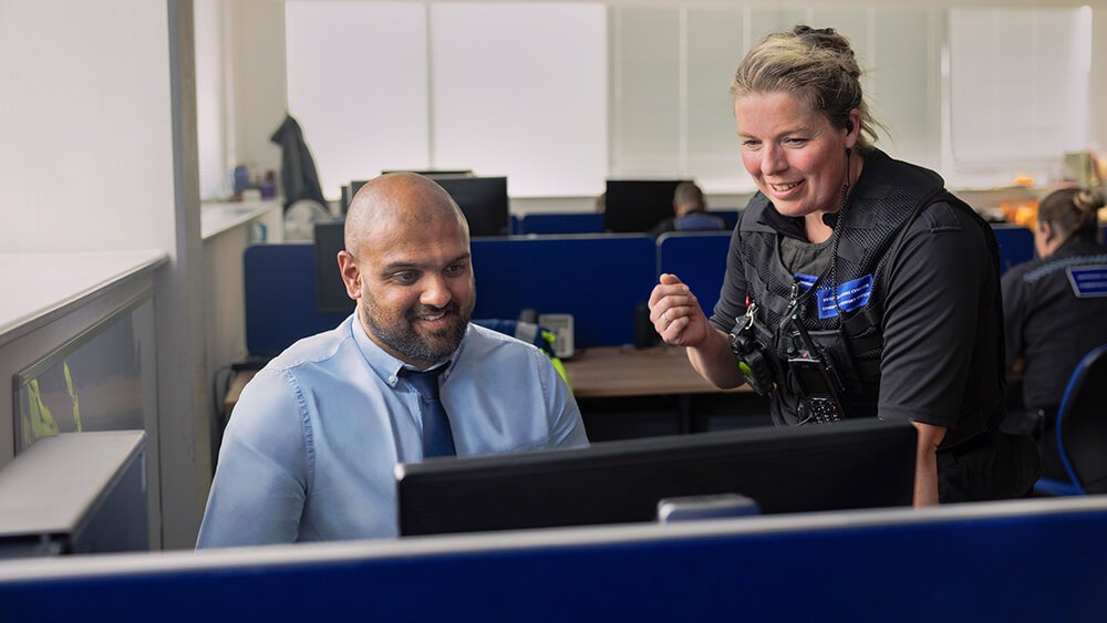 Male officer in plain clothes sitting at a desk with a uniformed female officer standing beside him.