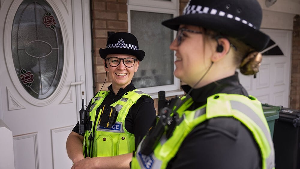 Two female officers in uniform, standing at the front door of a house. 